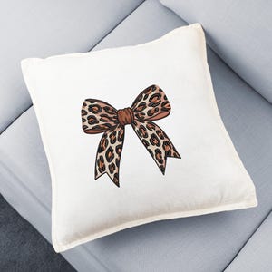 May include: A square, off-white decorative pillow featuring a large bow design. The bow has a leopard print pattern in shades of brown and black, with a darker brown center. The pillow is placed on a light blue sofa.