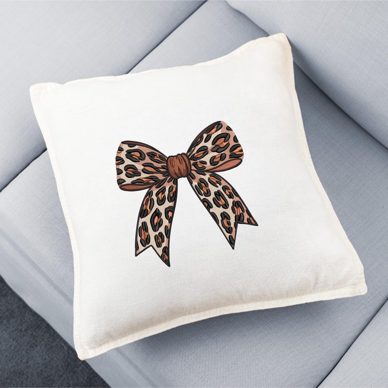 May include: A square, off-white decorative pillow featuring a large bow design. The bow has a leopard print pattern in shades of brown and black, with a darker brown center. The pillow is placed on a light blue sofa.