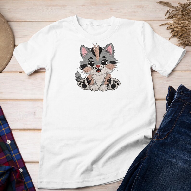 May include: White t-shirt featuring an embroidered cartoon kitten. The kitten has gray, brown, and pink fur, with large eyes and a smiling expression. The shirt is paired with blue denim jeans.