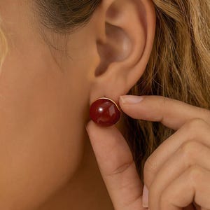 May include: A close-up of a red, round stud earring with a gold-colored border. The earring is a deep, rich red color and appears to be made of a smooth, polished material. The earring is being held by a person's fingers.
