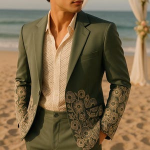 Op de afbeelding: Een saliegroen pak met bloemenborduurwerk op de mouwen en revers. Het pak wordt gecombineerd met een crèmekleurig overhemd met open weefsel. De setting is een strand, wat duidt op een bruiloft of speciale gelegenheid.