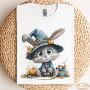 May include: A white short-sleeve t-shirt featuring a graphic illustration of a gray bunny wearing a blue witch hat. The bunny is surrounded by orange pumpkins and a small blue bucket labeled "WOCO" filled with colorful candies and treats.