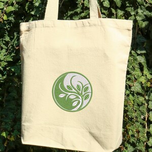 May include: A beige canvas tote bag with a green and white circular design featuring a stylized plant. The bag has two handles and is held up against a backdrop of green foliage.
