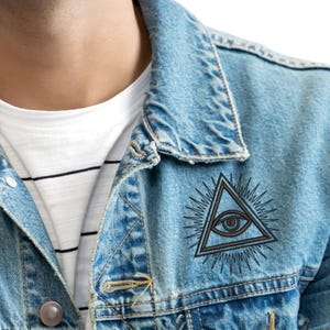 Peut inclure: Veste en jean bleu clair avec un motif d'œil dans un triangle noir et gris. La veste a une fermeture boutonnée, deux poches poitrine et un t-shirt rayé blanc et noir en dessous. Le motif est sur la poitrine supérieure droite.