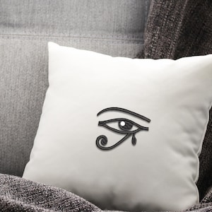 以下が含まれることがあります： 白い正方形のクッションに黒いホルスの目のデザイン。クッションは、灰色のテクスチャ加工された表面と濃い灰色の毛布の上に置かれています。ホルスの目は、古代エジプトの保護の象徴です。