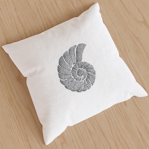 Könnte beinhalten: Ein quadratisches weißes Kissen mit einem grau gestickten Nautilus-Muschelmuster. Die Muschel ist ein detailliertes Spiralmuster, das auf dem Kissen zentriert ist. Das Kissen besteht aus einem strukturierten Stoff, vor hellem Holzgrund.