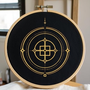 May include: A black embroidery hoop with a geometric gold design. The design features concentric circles, squares, and diamond shapes. The embroidery is set in a wooden hoop, creating a framed art piece.