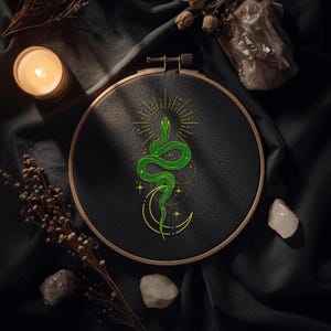 May include: An embroidery hoop with black fabric featuring a green snake design. The snake is coiled around a crescent moon with stars, and a sunburst design. The hoop is made of wood.