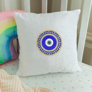 Peut inclure: Un coussin carré blanc avec un motif brodé d'œil protecteur bleu et or. Le coussin est en tissu doux et moelleux, posé sur un lit bébé blanc. Un coussin arc-en-ciel est partiellement visible en arrière-plan.