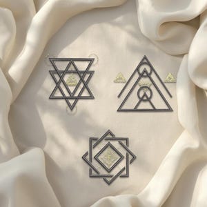 Puede incluir: Tres piezas decorativas geométricas, de color gris oscuro y dorado. Los diseños incluyen una estrella, un triángulo con un círculo y un cuadrado con un diamante. Cada pieza tiene un centro dorado y está sobre una tela crema.