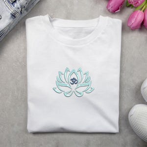 May include: White t-shirt with a light blue lotus flower design and an Om symbol. The lotus flower is embroidered on the front. The shirt is neatly folded. The design is a symbol of peace and spirituality.
