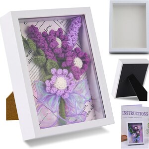 K&ouml;nnte beinhalten: Ein wei&szlig;er Schattenrahmen mit einem geh&auml;kelten Lavendelblumenarrangement. Die Blumen sind lila mit wei&szlig;en Zentren und gr&uuml;nen Bl&auml;ttern, akzentuiert durch ein Lavendelband. Der Rahmen hat einen St&auml;nder.
