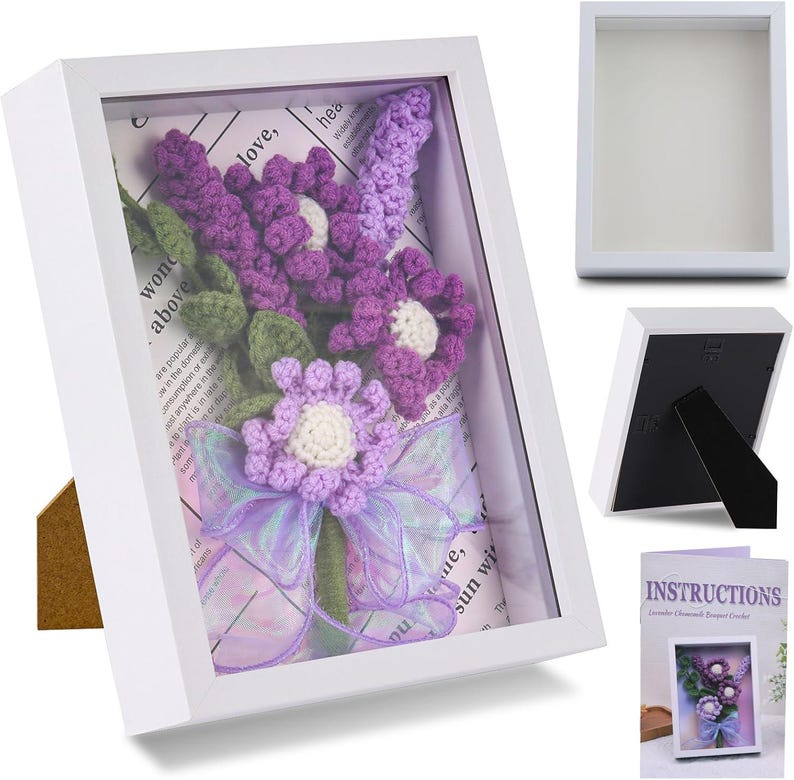 K&ouml;nnte beinhalten: Ein wei&szlig;er Schattenrahmen mit einem geh&auml;kelten Lavendelblumenarrangement. Die Blumen sind lila mit wei&szlig;en Zentren und gr&uuml;nen Bl&auml;ttern, akzentuiert durch ein Lavendelband. Der Rahmen hat einen St&auml;nder.
