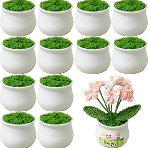 May include: Twelve white ceramic pots, eleven filled with green faux moss and one containing a crocheted pink flower arrangement. The flower pot features a painted label with a heart, a tulip, and the text "I love you" in black lettering.