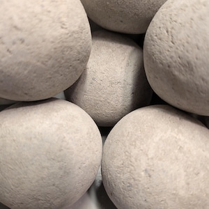 May include: Close-up of several smooth, light grey stones. The stones are round and have a slightly textured surface. The stones are piled together, creating a natural, organic aesthetic. The colour is a muted, neutral tone.