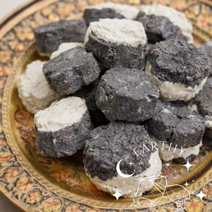 May include: A collection of dark gray and white, round, textured objects arranged on a decorative gold-colored plate. The objects have a layered appearance, with a dark gray base and a white top. The plate has an ornate, embossed design and the text "EARTHY CRUNCH".