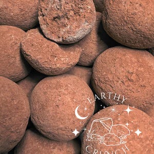 Può includere: Primo piano di un mucchio di oggetti rotondi marrone-rossastri con una superficie ruvida e strutturata. Alcuni oggetti sono interi, mentre altri sono rotti, rivelando un interno simile. L'immagine include il testo "EARTHY CRUNCH" con una grafica a mezzaluna e stella.