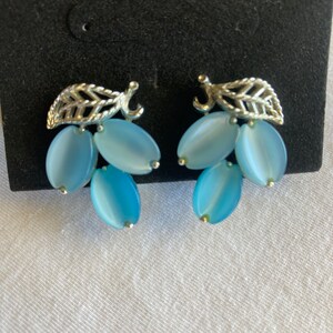May include: A pair of vintage earrings featuring a silver-toned leaf design at the top, with three light blue oval-shaped beads hanging below. The earrings are set against a black background.