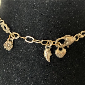 May include: Silver necklace featuring a floral charm, a leaf charm, and a heart charm. The necklace is set against a black backdrop, highlighting the delicate chain and the charms' details.