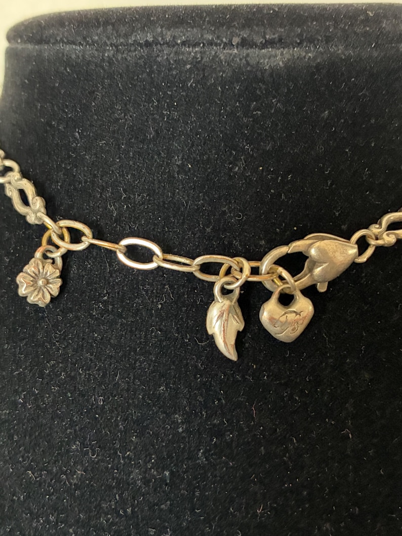 May include: Silver necklace featuring a floral charm, a leaf charm, and a heart charm. The necklace is set against a black backdrop, highlighting the delicate chain and the charms' details.