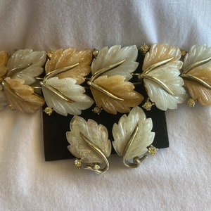 May include: A vintage jewelry set featuring a bracelet and matching earrings. The bracelet and earrings are designed with leaf-shaped links in shades of cream and tan, accented with silver-tone metal and small clear rhinestones. The set is displayed on a black surface.