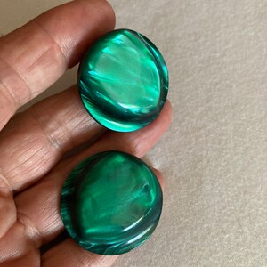 May include: Two round, iridescent green buttons with a swirling pattern. The buttons have a glossy finish and a slightly domed shape, reflecting light. These buttons could be used for clothing or craft projects.