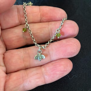 May include: A silver chain bracelet featuring a bee charm and three small green gemstone beads. The bee pendant is detailed, and the bracelet is delicate. The gemstones are small and round.