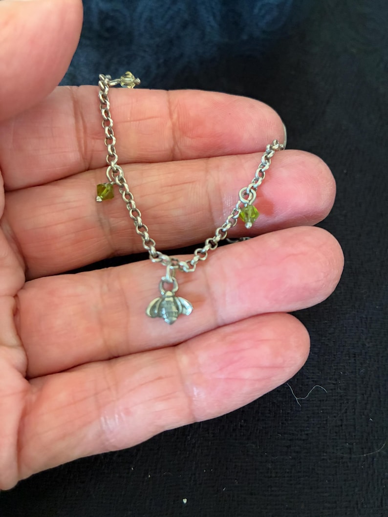 May include: A silver chain bracelet featuring a bee charm and three small green gemstone beads. The bee pendant is detailed, and the bracelet is delicate. The gemstones are small and round.