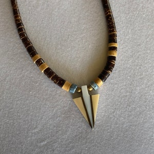 May include: A necklace featuring dark brown, round beads with gold-colored spacers. The pendant is a triangle shape with a cream-colored center and gold-colored edges. The necklace is displayed against a gray background.