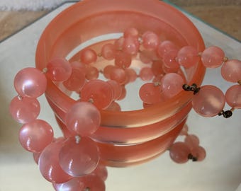 May include: A translucent peach-colored bangle bracelet paired with a matching beaded necklace. The necklace consists of round, smooth, semi-translucent peach beads strung together, resting partially inside the circular bangle on a reflective surface.