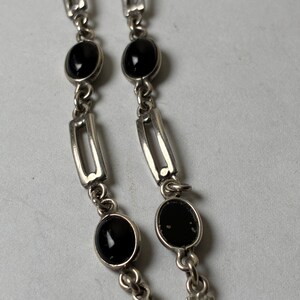 Puede incluir: Pulsera plateada con piedras negras ovaladas. La pulsera tiene una serie de segmentos enlazados, con las piedras engastadas entre eslabones metálicos rectangulares y curvos. Se ve un cierre al final de la pulsera.