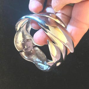 May include: Silver-toned metal cuff bracelet with a braided design. The bracelet has a hinged clasp and a polished finish. The design features overlapping, curved elements that create a textured appearance. The bracelet is shown against a dark background.