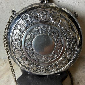 May include: A round, silver-toned metal compact purse featuring an intricate embossed floral and scrollwork pattern surrounding a smooth circular center. The item includes a hinged clasp closure at the top and a thin, attached metal chain strap.