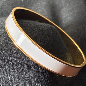 Puede incluir: Un brazalete dorado con una ancha banda de esmalte blanco. El brazalete tiene un diseño sencillo y elegante, con un acabado liso y pulido. La banda blanca está bordeada por finas líneas doradas.