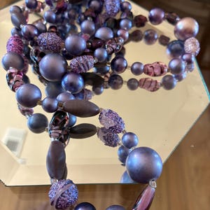 Puede incluir: Un collar vintage con una variedad de cuentas moradas de diferentes formas y tamaños. El collar incluye cuentas redondas, ovaladas y texturizadas en tonos lavanda, amatista y ciruela. El collar se exhibe sobre una superficie espejada.