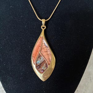 May include: A gold-toned pendant necklace with a teardrop-shaped pendant. The pendant features a mosaic of peach and brown tones, with iridescent accents. The necklace is displayed on a black surface.