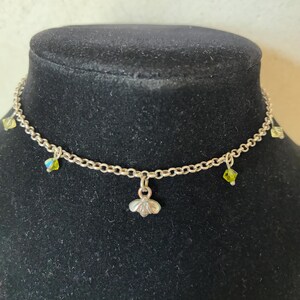 May include: A delicate silver necklace with a small, flower-shaped pendant. The necklace features small, sparkling green and yellow crystals. The necklace is displayed on a black mannequin.