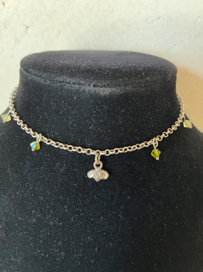 May include: A delicate silver necklace with a small, flower-shaped pendant. The necklace features small, sparkling green and yellow crystals. The necklace is displayed on a black mannequin.