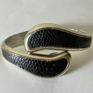May include: A silver-toned cuff bracelet with black textured accents. The bracelet features a curved design with a silver border around the black textured sections. The bracelet is shown against a white background.