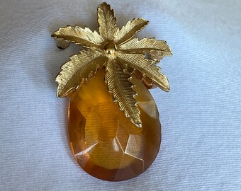 Op de afbeelding: Goudkleurige broche met een gestileerd ananasontwerp. De bovenkant van de broche heeft een gedetailleerd palmbladontwerp en het lichaam is een gefacetteerde, amberkleurige steen. Deze vintage broche is geschikt voor een vleugje elegantie.