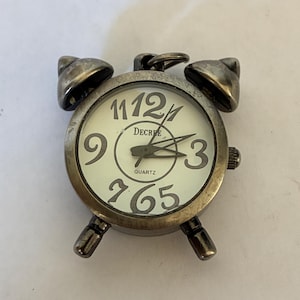 May include: A small, round, metallic alarm clock pendant with a weathered bronze finish. The off-white clock face features large, dark, stylized numerals, the word DECREE, and the word QUARTZ. It includes two top bells and a small hanging loop.