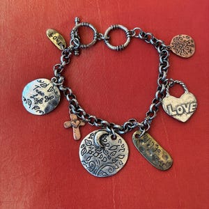 Op de afbeelding: Zilverkleurige bedelarmband met diverse hangers. Bedels zijn onder meer een kruis, een hart met "LOVE", een levensboom en andere gegraveerde labels. De armband ligt op een rode ondergrond.