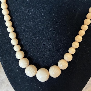 May include: Cream-colored beaded necklace. The necklace features round beads of varying sizes, with the larger beads having a textured, ridged surface. The beads are strung together, creating a classic jewelry piece.