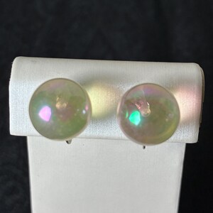 May include: A pair of iridescent clip-on earrings. Each earring is a round, semi-transparent sphere with a pearlescent sheen, reflecting hues of pink, green, and yellow. The earrings are mounted on a white display card against a dark background.