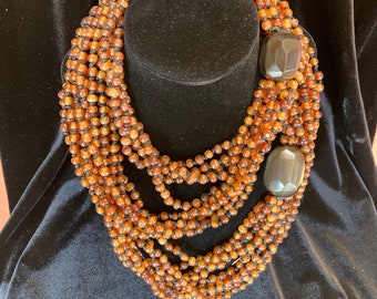 Peut inclure: Un collier à plusieurs rangs composé de petites perles rondes de couleur ambre enfilées sur un fil noir. Le collier comporte deux grands accents sombres de forme géométrique. Le collier est exposé sur un mannequin noir.