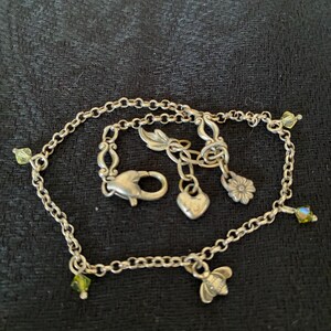 May include: Silver chain bracelet with various charms. Charms include a heart, a flower, and a bee. The bracelet also features small green and iridescent crystal accents. The clasp is a lobster claw style.