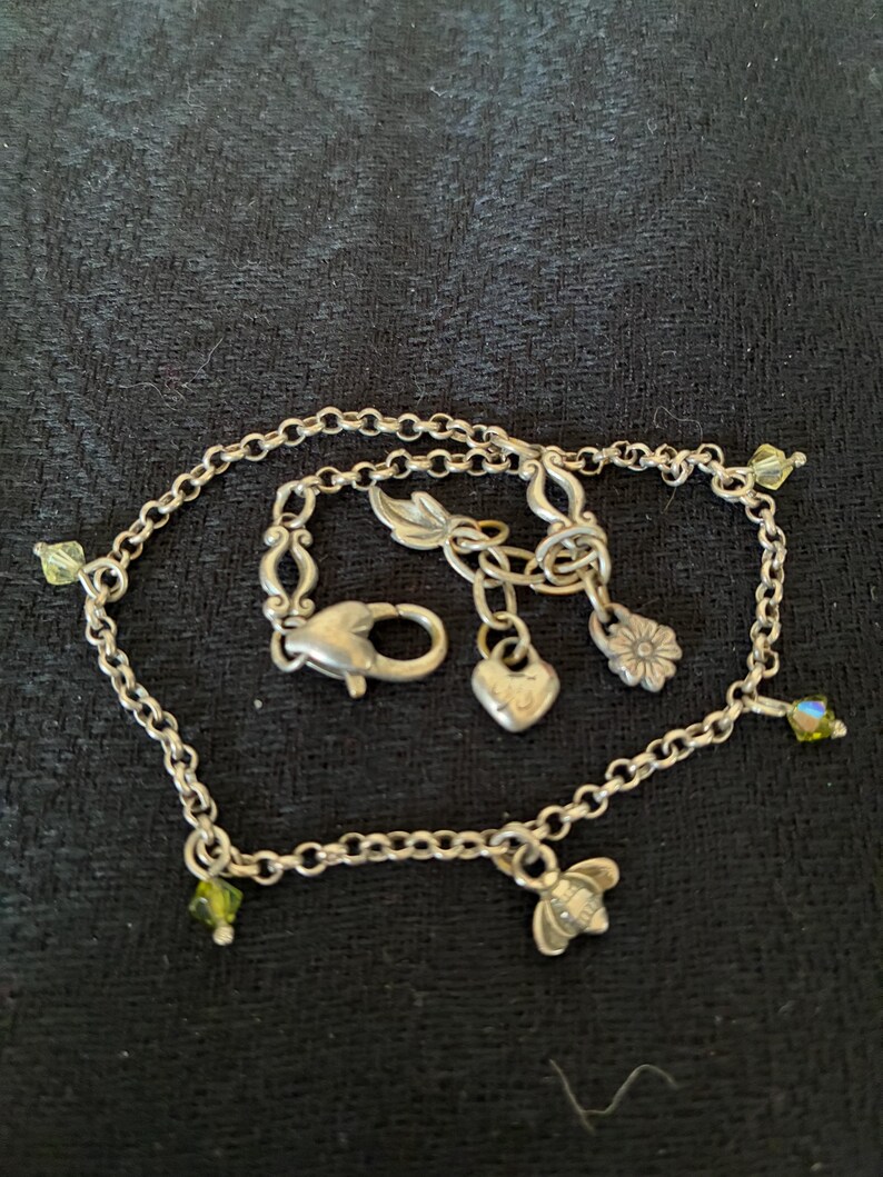 May include: Silver chain bracelet with various charms. Charms include a heart, a flower, and a bee. The bracelet also features small green and iridescent crystal accents. The clasp is a lobster claw style.