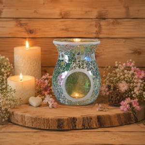 Könnte beinhalten: Ein Mosaikglas-Ölwärmer mit grün-weißem Kachelmuster und einer kreisförmigen Öffnung. Der Wärmer ist umgeben von zwei weißen Stumpenkerzen, kleinen rosa Blüten und einem herzförmigen Stein auf einer runden Holzscheibe als Basis.