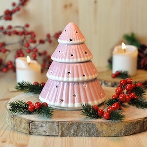 May include: A pink ceramic Christmas tree with star-shaped cutouts and white trim. The tree is on a wooden slice with red berries and lit candles in the background. Festive holiday decor.