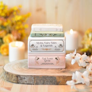 Könnte beinhalten: Ein Stapel dekorativer Keramikbücher in Weiß, Grün und Rosa mit goldenen Akzenten. Das mittlere Buch trägt die Aufschrift "Myths, Fairy Tales & Legends" mit Schwanenillustrationen. Das untere Buch trägt die Aufschrift "Let Love Grow".